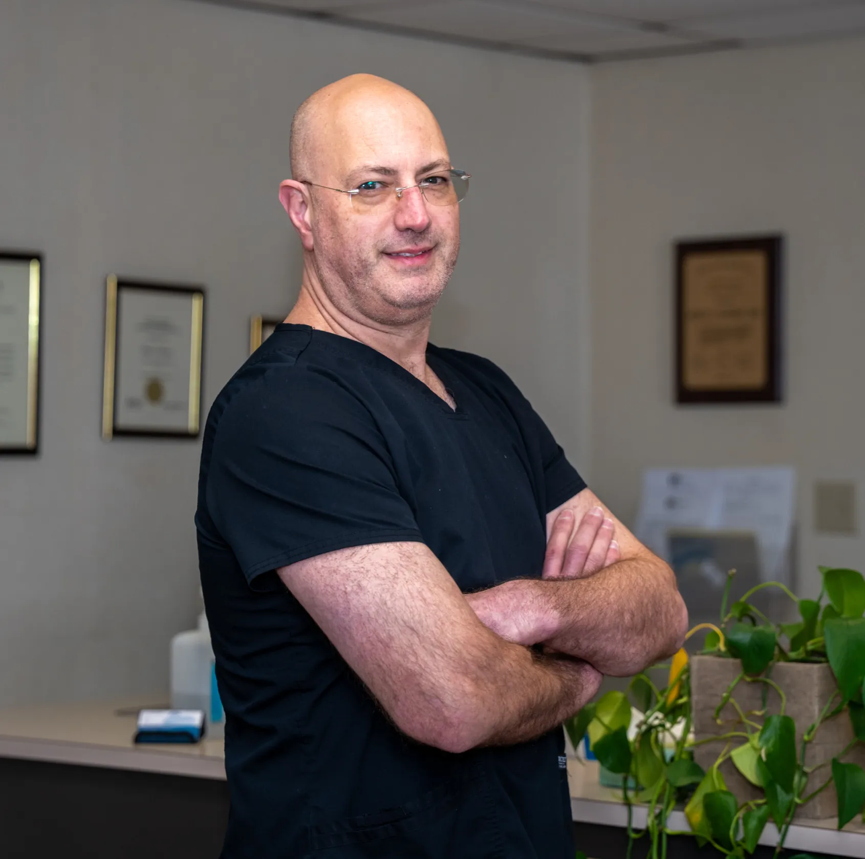 Bald man in glasses wearing black medical scrubs with arms crossed in an office setting.