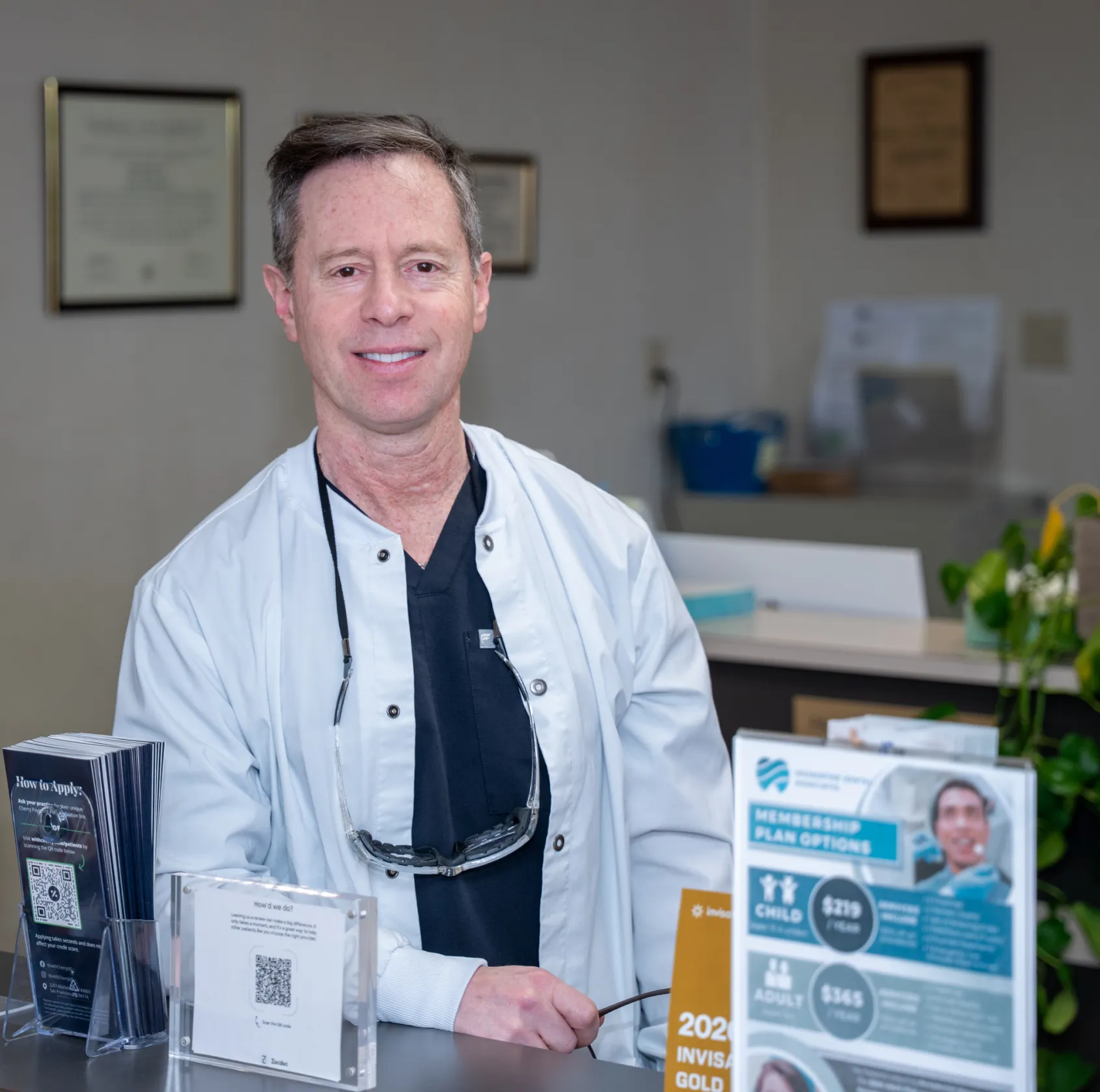 Smiling male dentist in white coat and black scrubs behind a clinic reception desk with informational brochures.