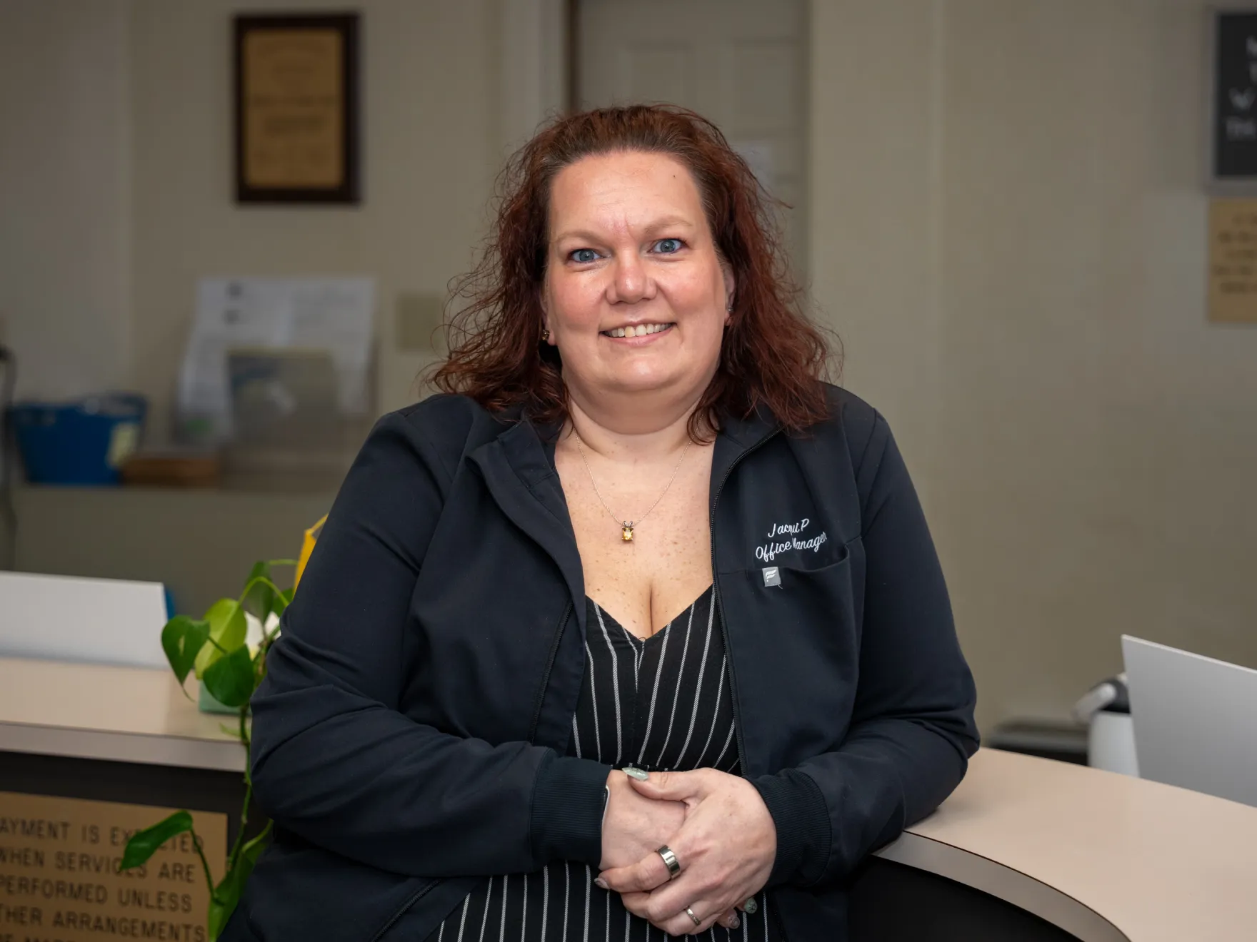 Smiling woman with curly hair wearing a black jacket labeled Office Manager, sitting at a reception desk.