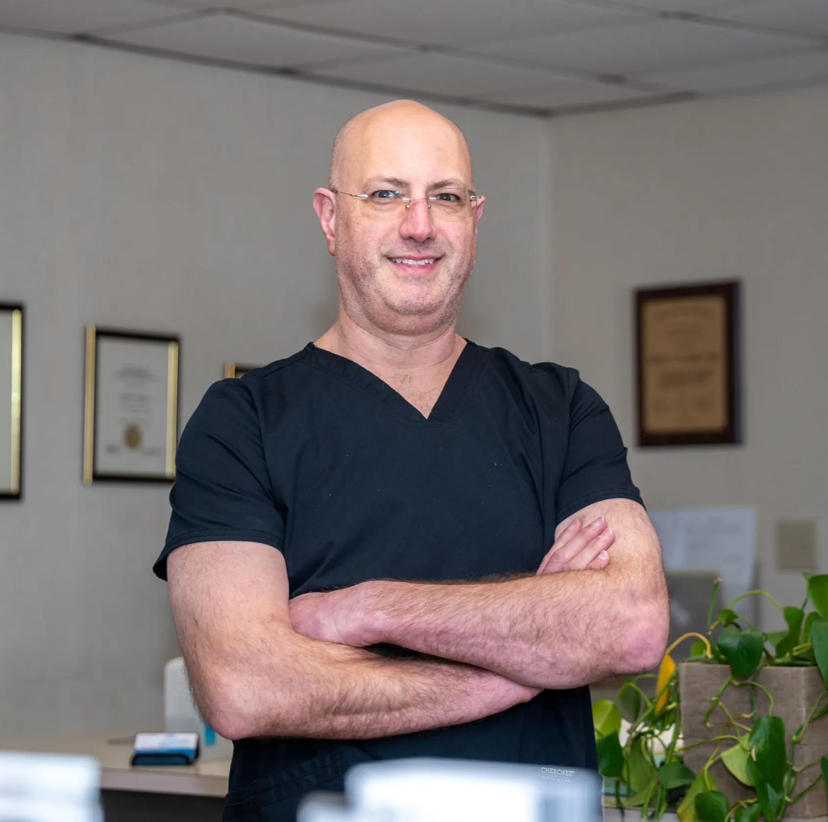 Smiling bald man in black medical scrubs with glasses stands with arms crossed indoors.