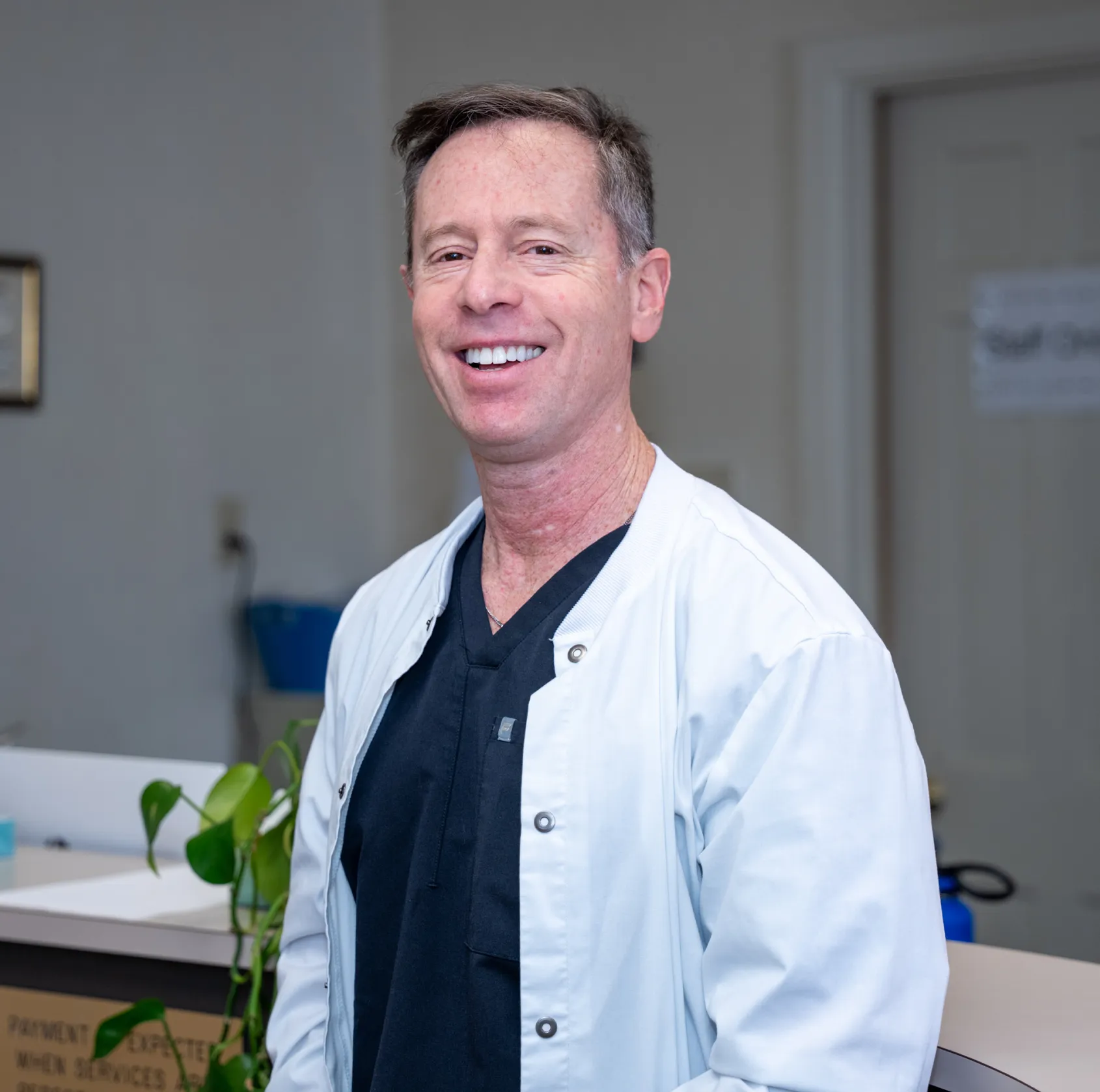 Smiling male medical professional wearing black scrubs and a white lab coat in a clinic setting.