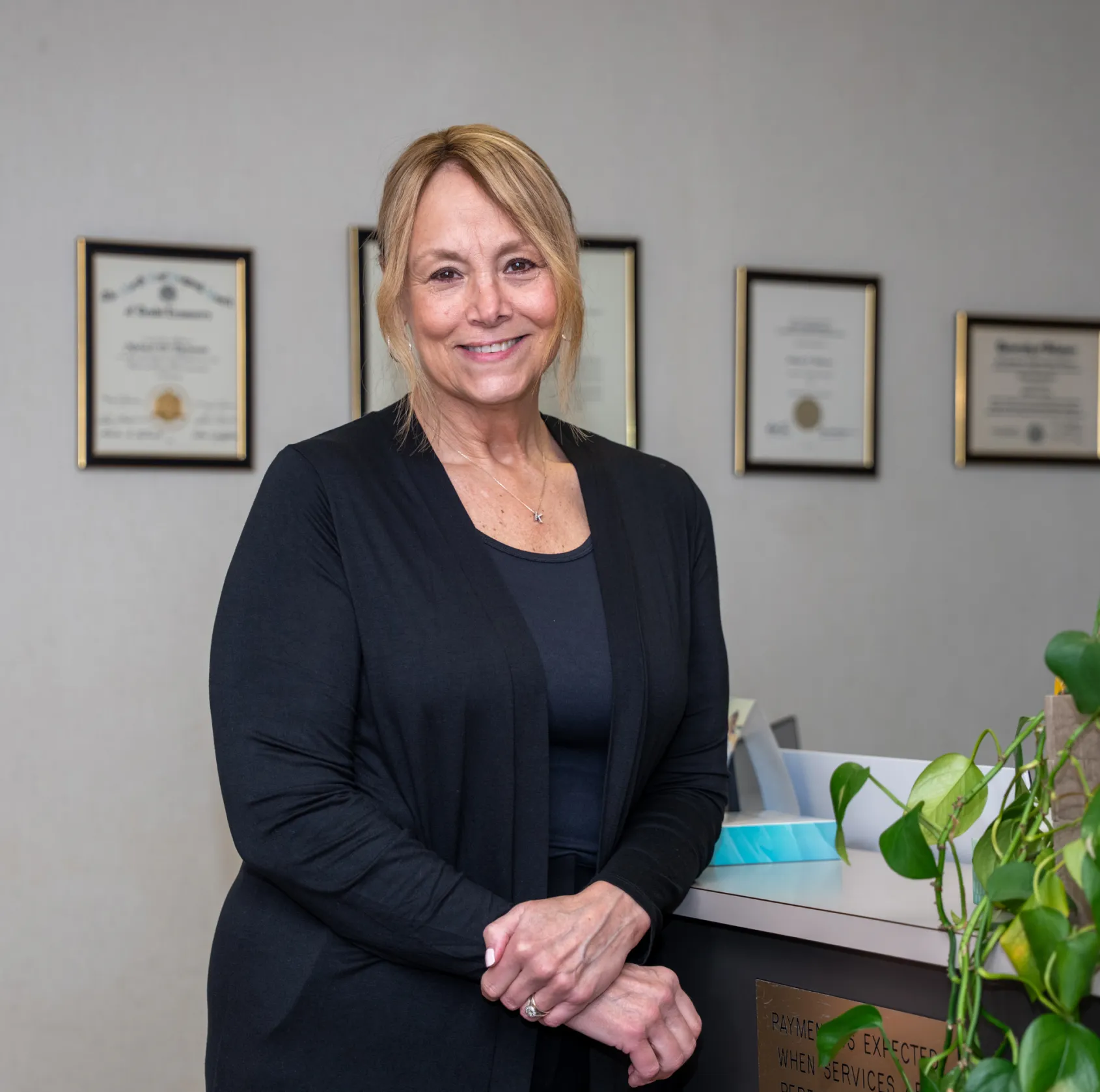 Smiling middle-aged woman in black stands in office with certificates on wall and plants nearby.
