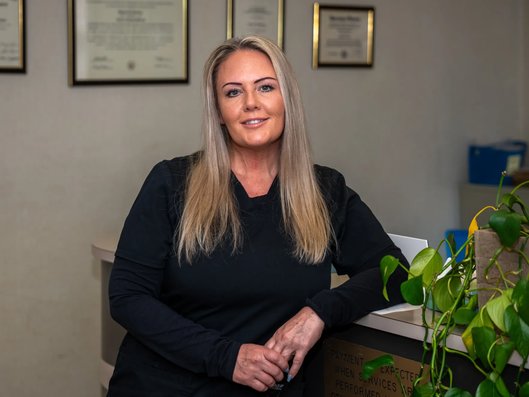 Blonde woman in black scrubs leaning on a desk with framed certificates in the background.