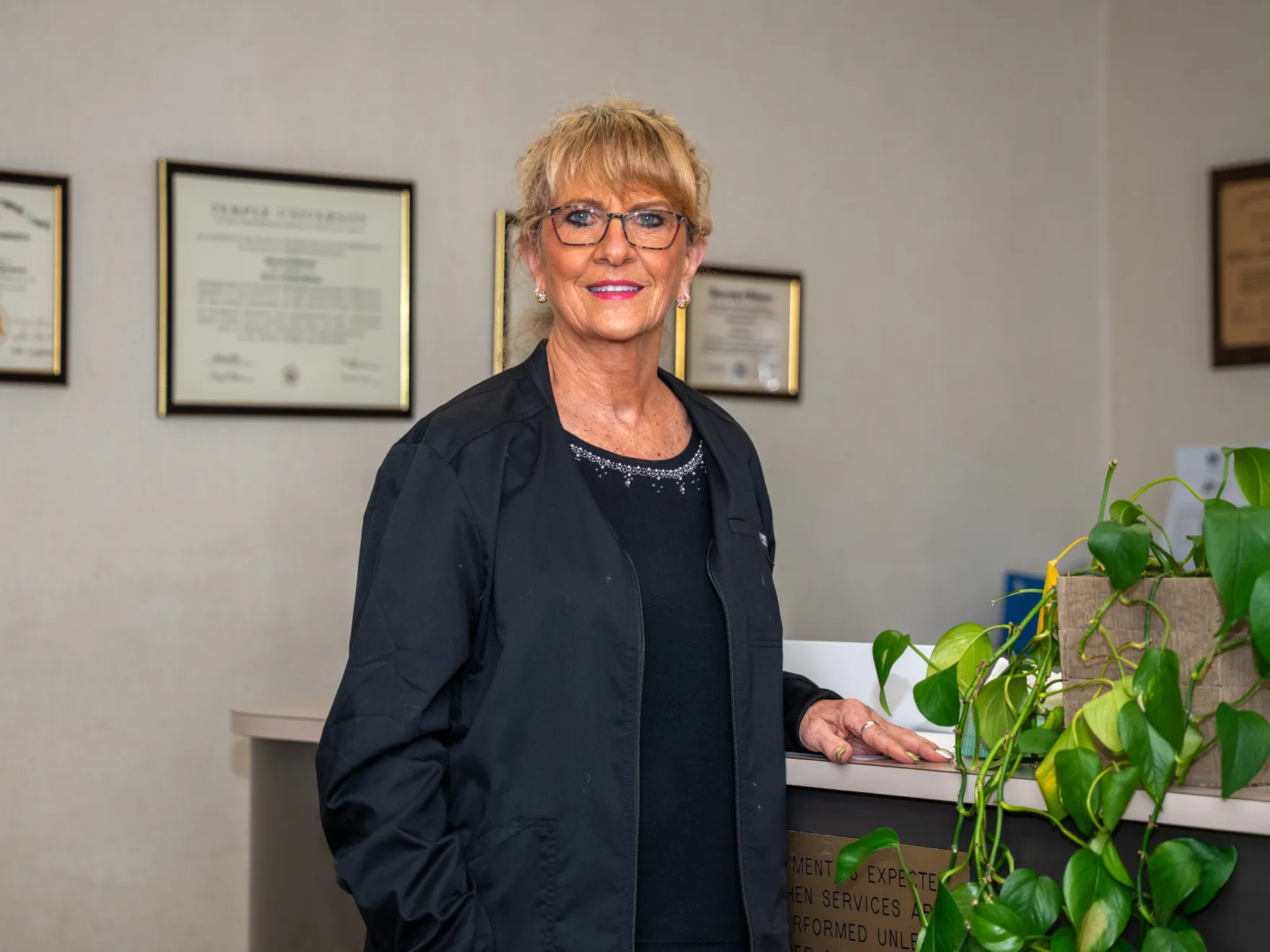 Smiling woman in glasses and black jacket standing by a desk with framed certificates and a plant nearby.