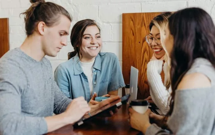 Group of four young adults smiling and using a tablet while sitting at a wooden table in a casual indoor setting.