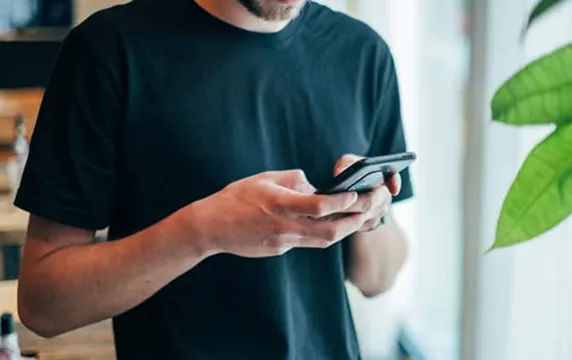 Person in black t-shirt holding and using a smartphone near a window with green plant leaves visible.