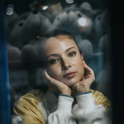 Young woman resting her chin on her hands, looking thoughtfully through a window with blurred teddy bears in the background.