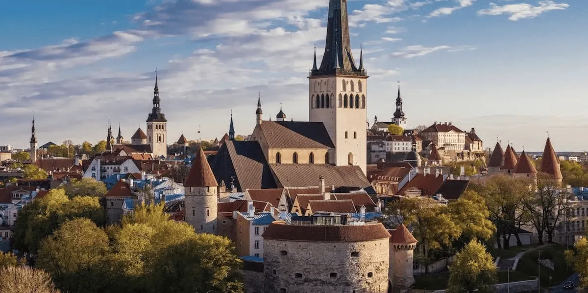 Panoramic view of a historic European city with medieval stone towers and a large church with a tall spire under a partly cloudy sky.