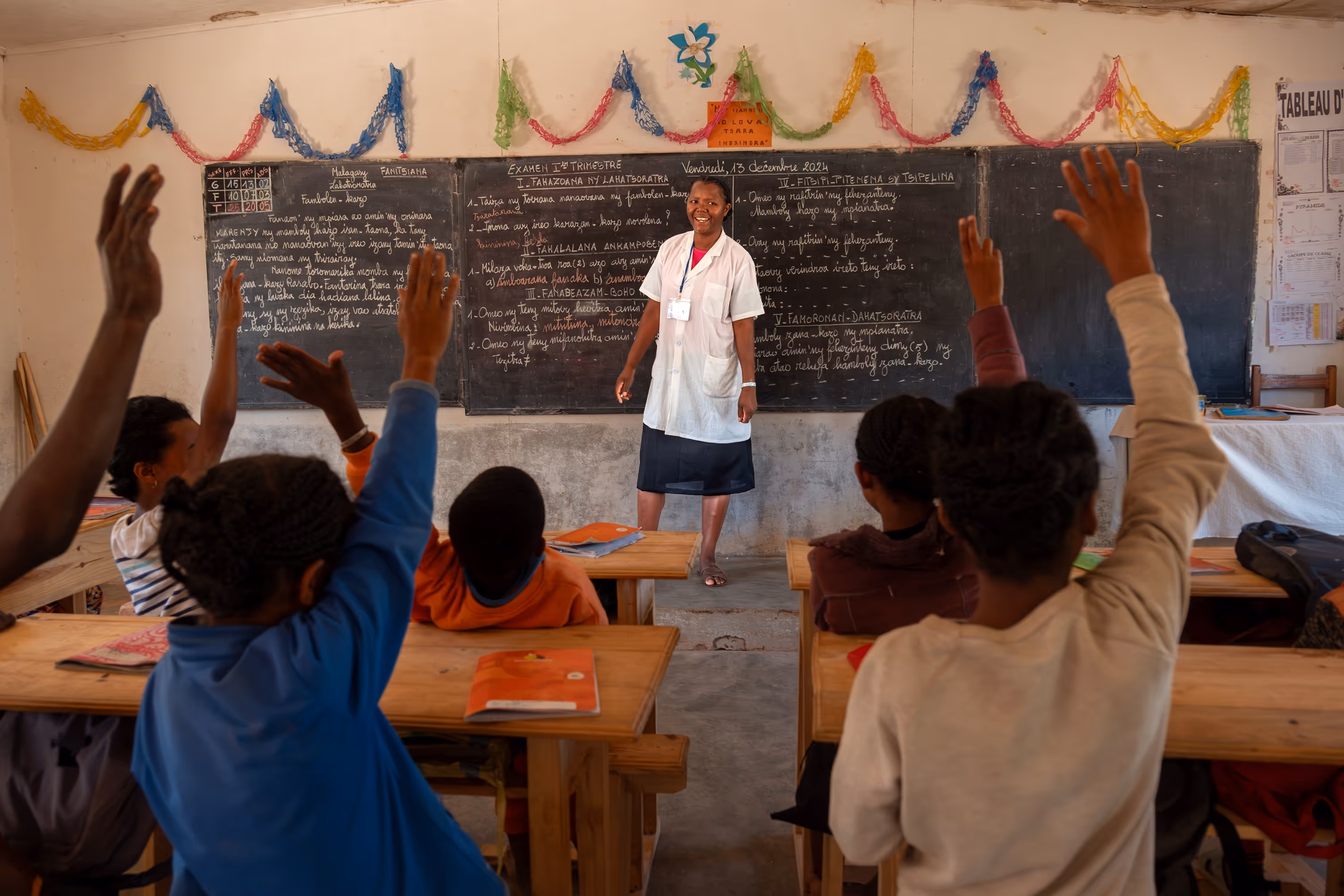 Classroom with teacher in front of a blackboard and children with their hands raised.