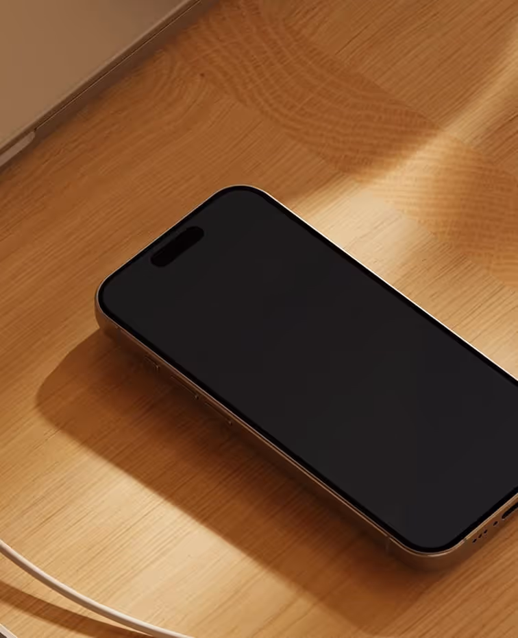 Mobilní telefon s černou obrazovkou ležící na dřevěném stole vedle bílého kabelu.