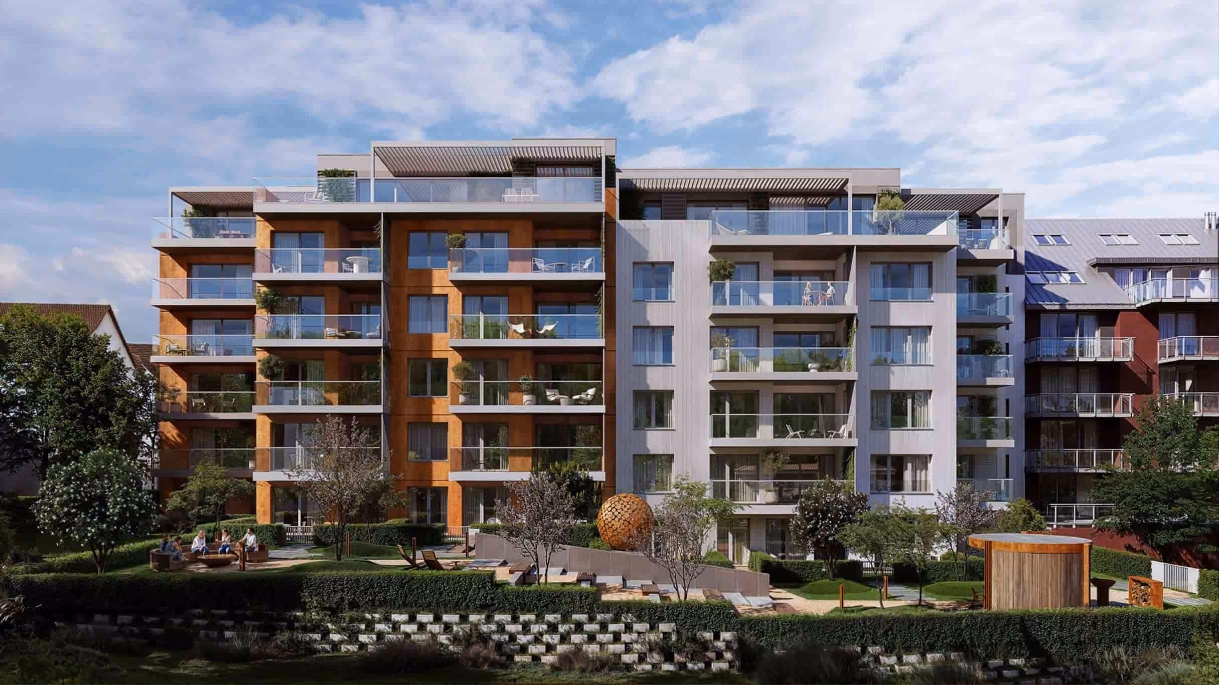 Moderní bytový komplex s několika patry a balkony, před ním trávník s posezením a zahradními rostlinami, pod oblohou s rozptýlenými mraky.