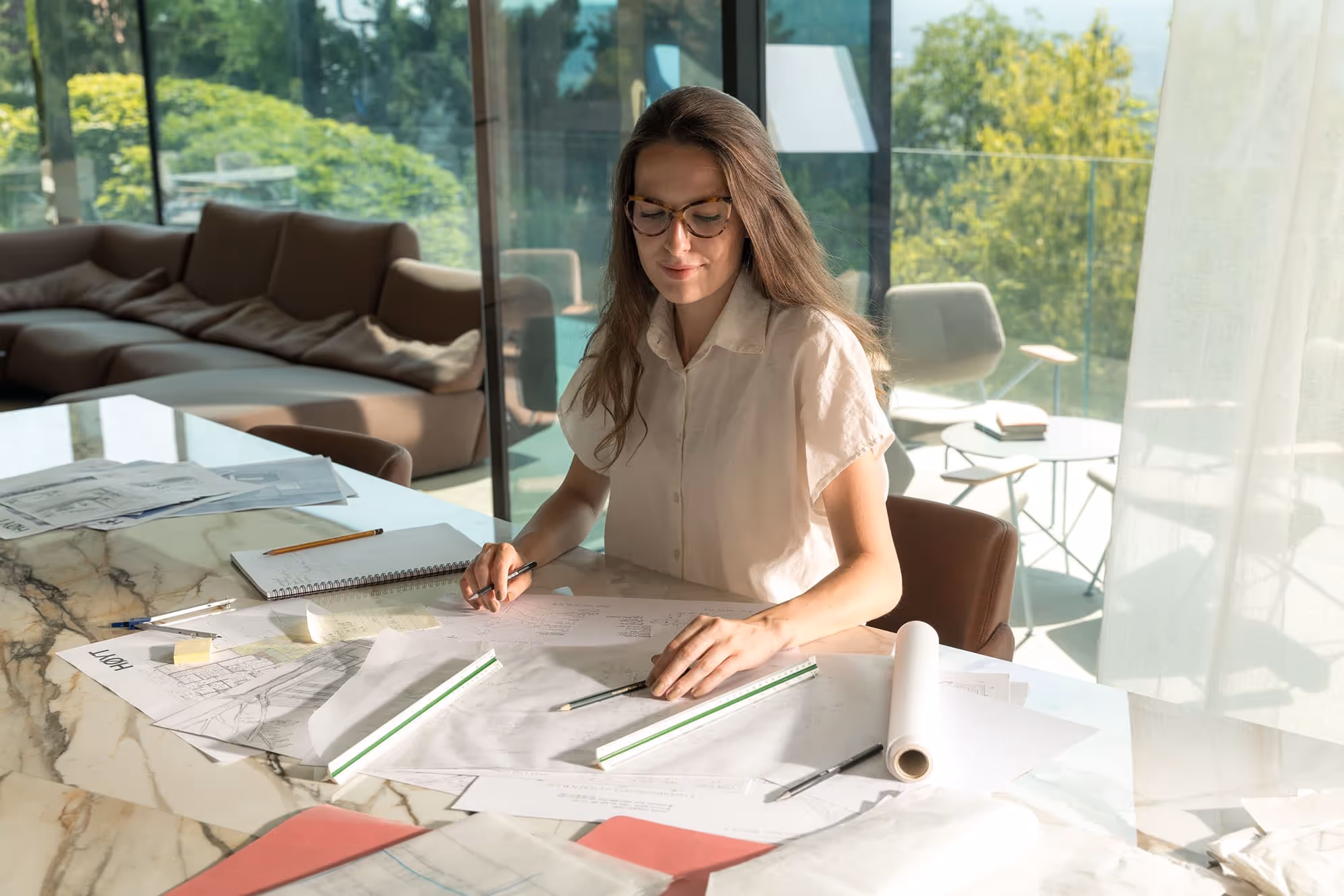 Žena v brýlích pracující u stolu s architektonickými plány a náčrty v moderní světlé místnosti s výhledem na zeleň.
