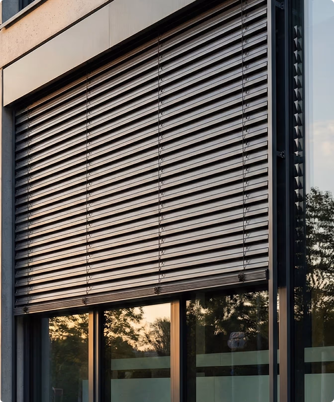 Venkovní hliníkové žaluzie na velkém okně moderní budovy s odrazem stromů a západu slunce.