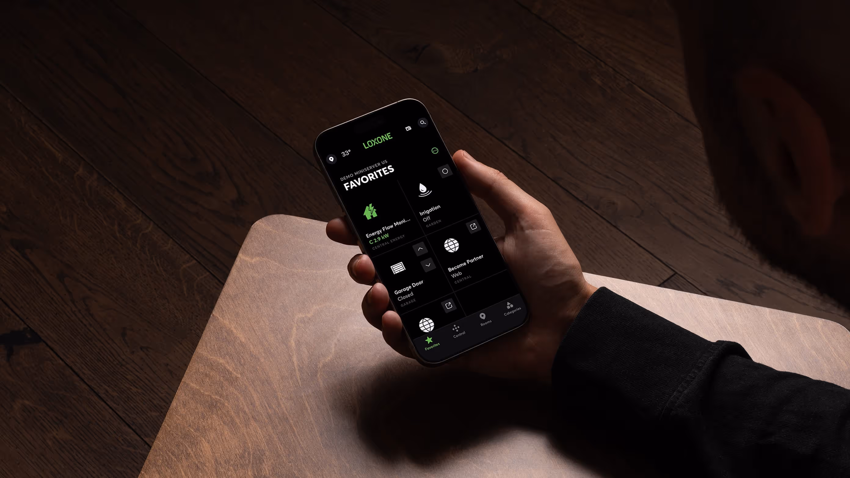 Osoba drží chytrý telefon s aplikací Loxone zobrazující oblíbené funkce domácí automatizace na černém pozadí.