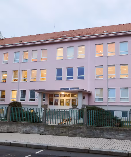 Fasáda růžové třípatrové školní budovy s osvětlenými okny a zeleným plotem před vchodem.