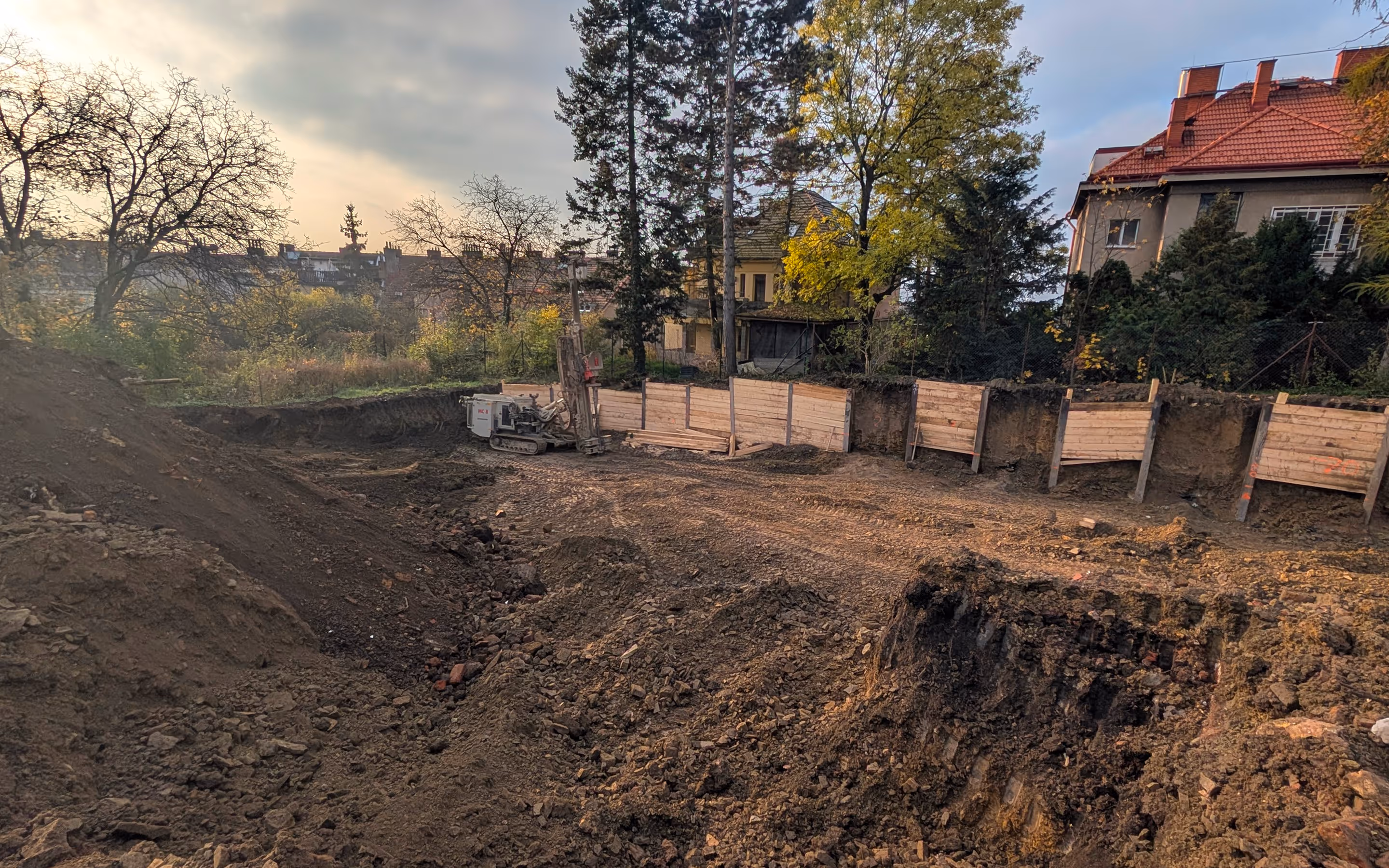 Stavební výkop na zasypaném pozemku s dřevěnou zídkou a stavební technikou za podzimního dne.