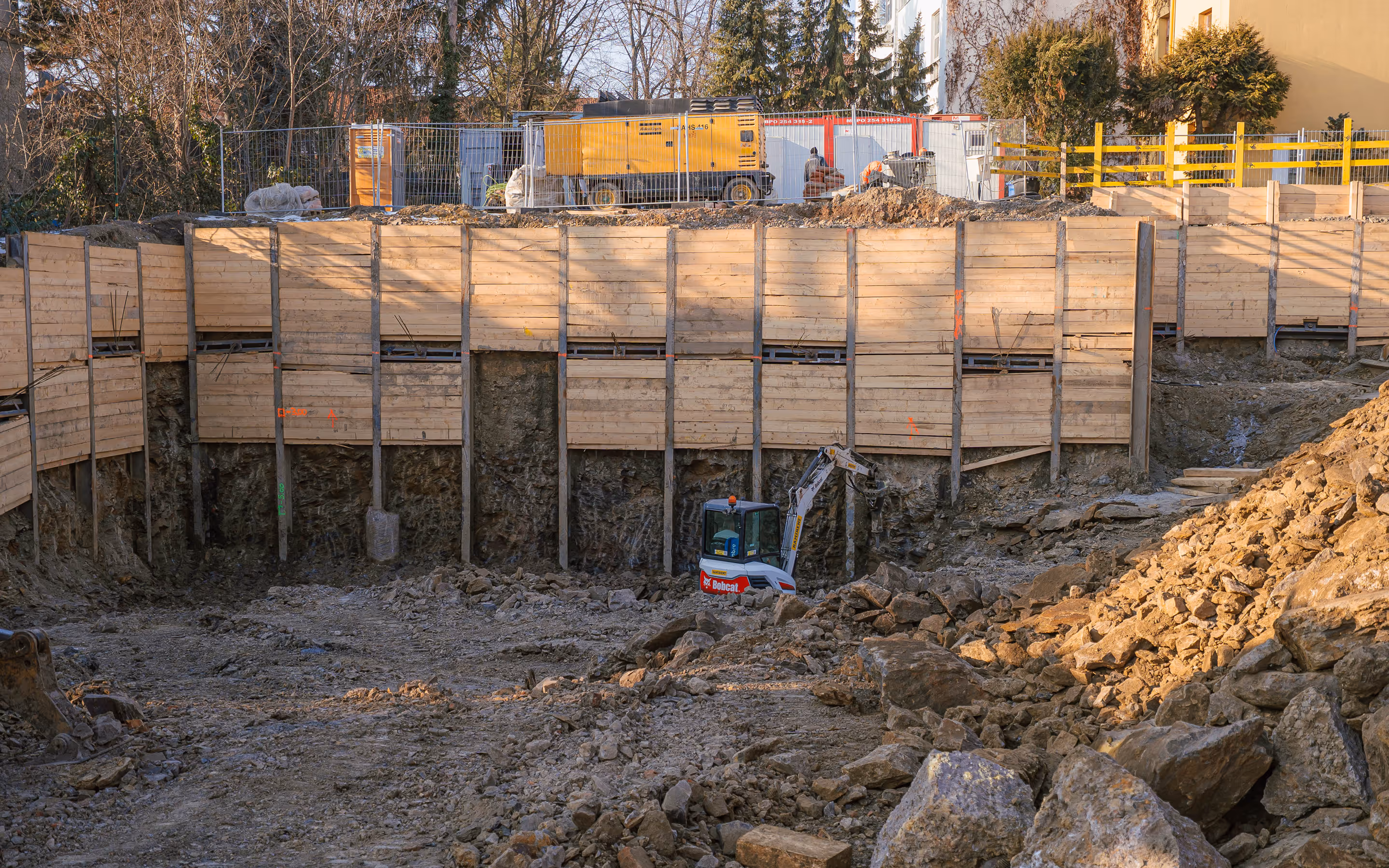 Stavební výkop na zasypaném pozemku s dřevěnou zídkou a stavební technikou za podzimního dne.