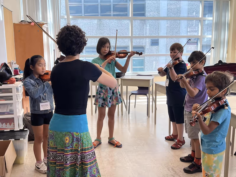 Music teacher instructing a group of five children playing violins in a bright classroom with large windows.
