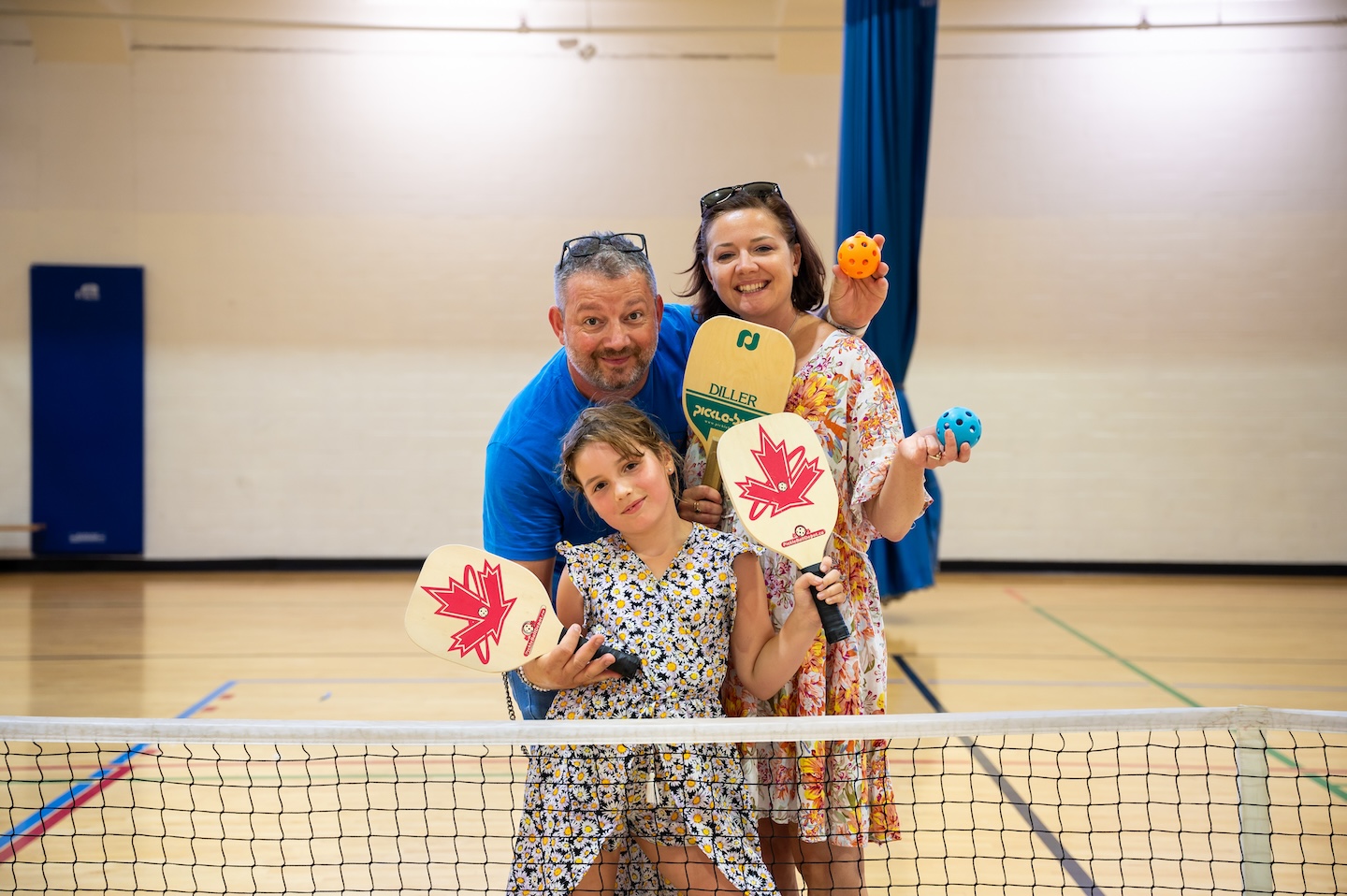 Smiling family of three holding pickleball paddles and balls on an indoor court behind the net.