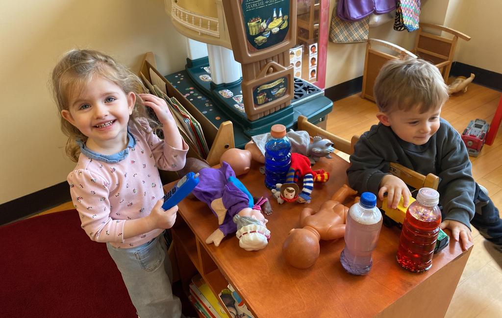 Two toddlers playing at a wooden table with dolls, toy trucks, and plastic bottles in a playroom.