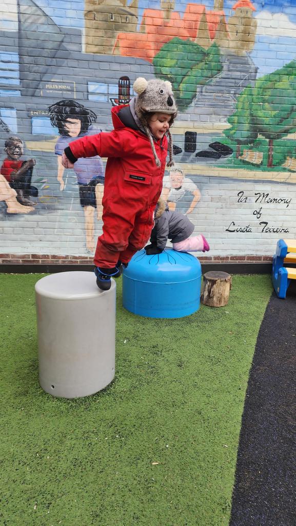 Child in a red snowsuit and animal hat jumping from a gray cylindrical stool to a blue one on artificial grass with a mural in the background.