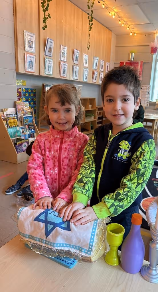 Two young children smiling and standing at a table in a classroom, with their hands on a covered loaf of challah bread decorated with a Star of David.