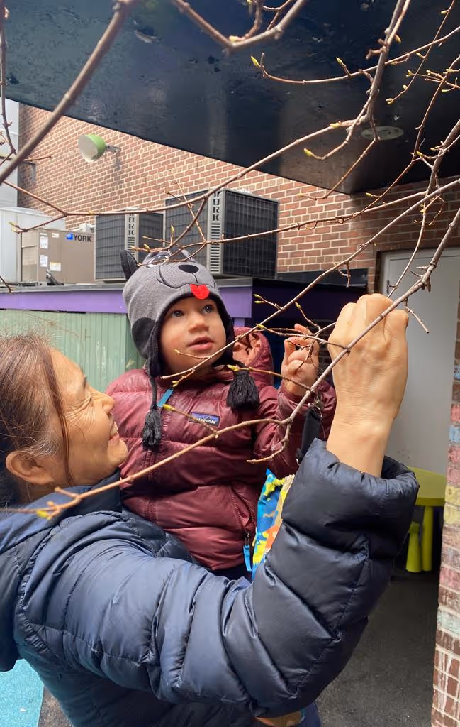 A woman holding a toddler wearing a gray animal hat with ears and a face, both reaching for small buds on tree branches.