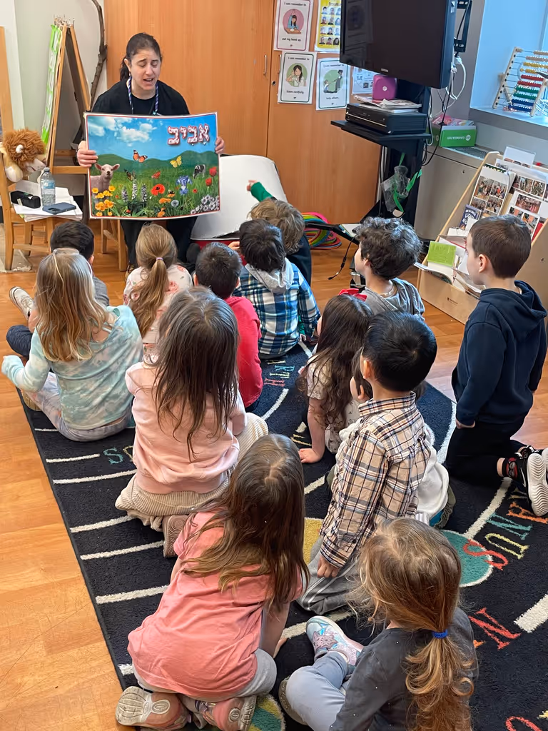 Teacher sitting on a chair holding a colorful nature poster while a group of young children sit on a carpet attentively watching her in a classroom.