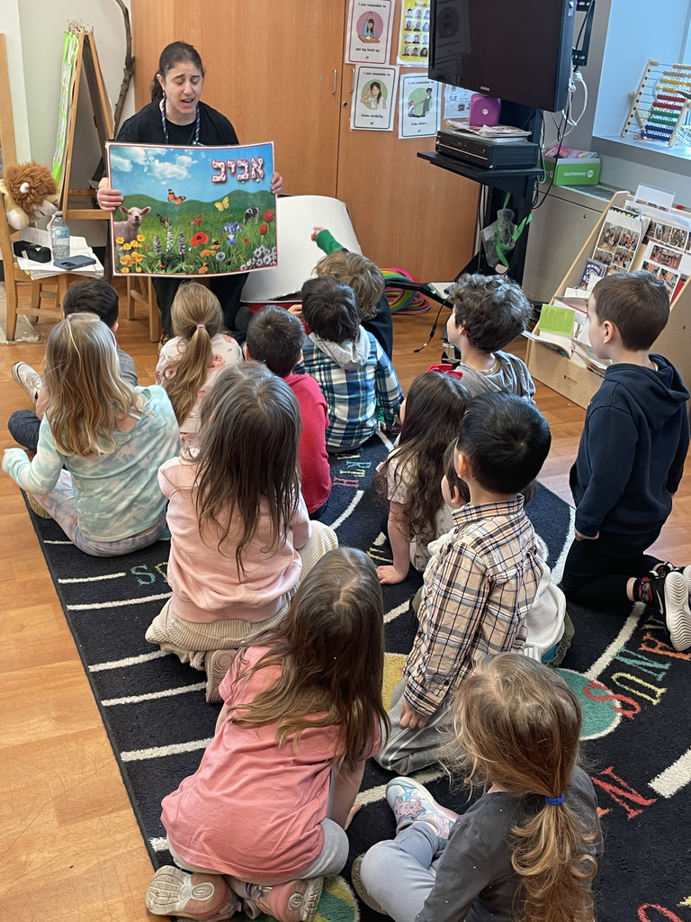 Teacher sitting on a chair holding a colorful nature poster while a group of young children sit on a carpet attentively watching her in a classroom.