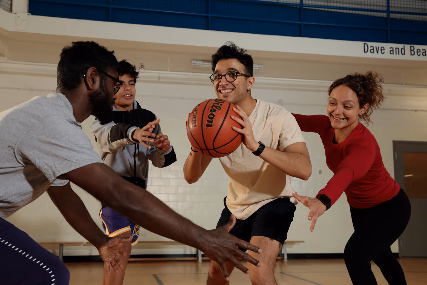 Four people playing basketball indoors with one person holding the ball and others reaching to block it.