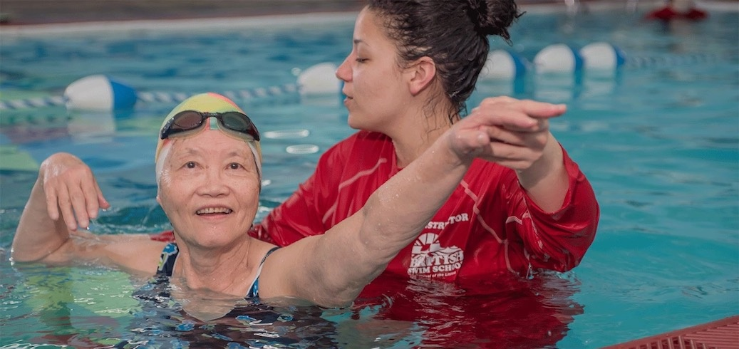 Swimming instructor in red shirt assists an elderly woman wearing a swim cap and goggles in a pool.