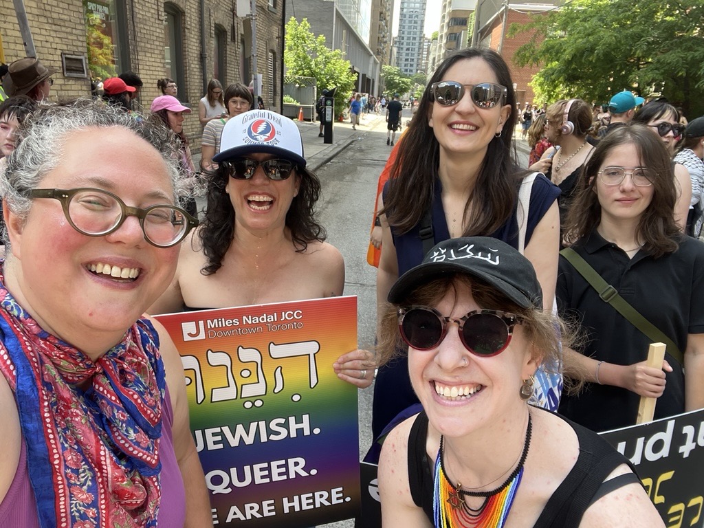 Five smiling people at an outdoor pride march, one holding a rainbow sign with the words 'Jewish. Queer. We are here.'