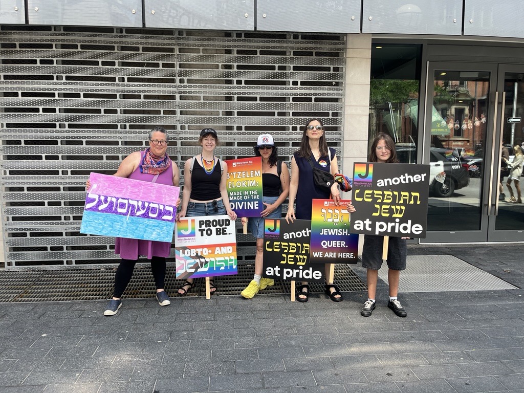 Five people standing outdoors holding colorful signs with LGBTQ+ and Jewish pride messages in English and Hebrew.