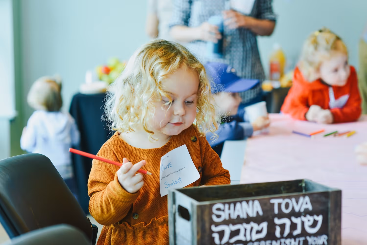 Young girl with curly blonde hair holding a red pencil and looking into a wooden box with Hebrew text saying 'Shana Tova' during a group activity.