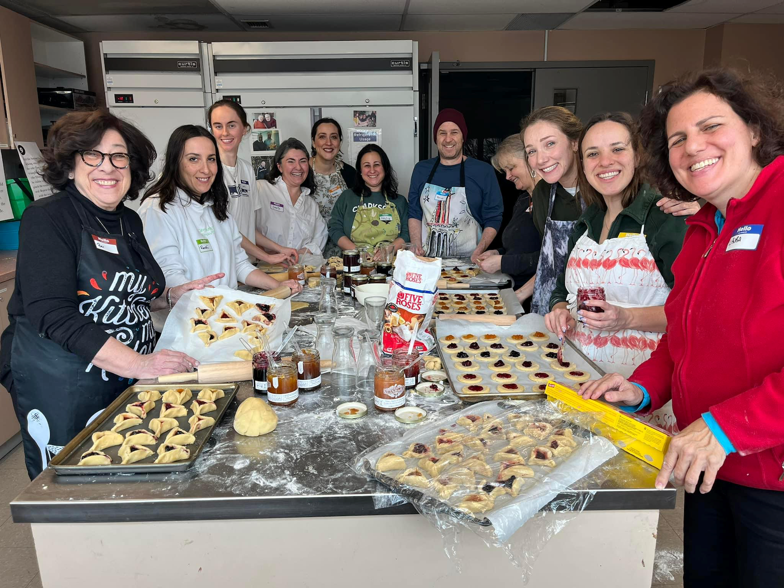 Group of people smiling and preparing filled pastries in a kitchen, with trays, rolling pins, jars of jam, and dough on the countertop.