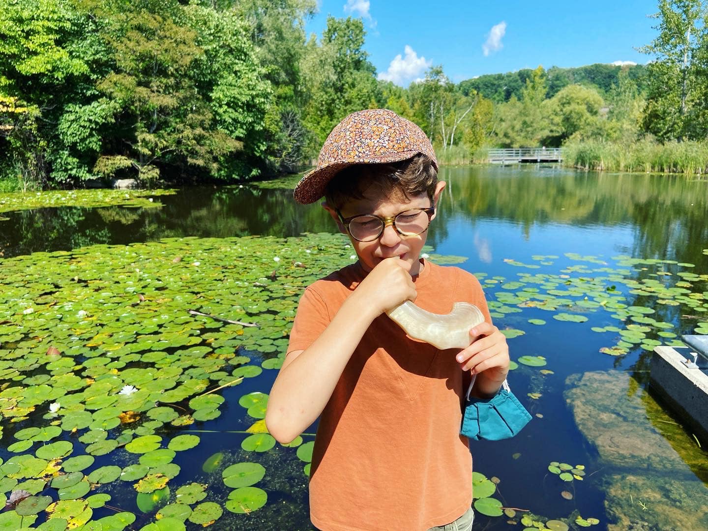Boy wearing glasses and a floral cap blowing a shofar horn by a pond with lily pads under a clear blue sky.