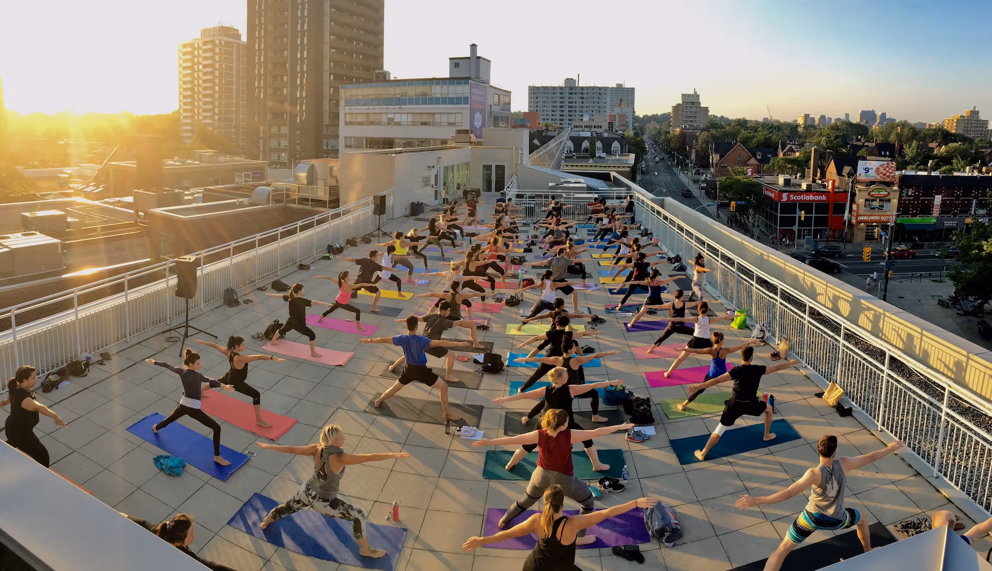 Large group of people practicing yoga on colorful mats on a rooftop at sunset in an urban setting.