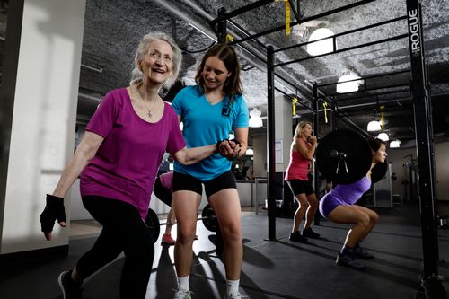 Older woman in purple workout clothes assisted by a trainer in blue during exercise in a gym with others lifting weights in the background.