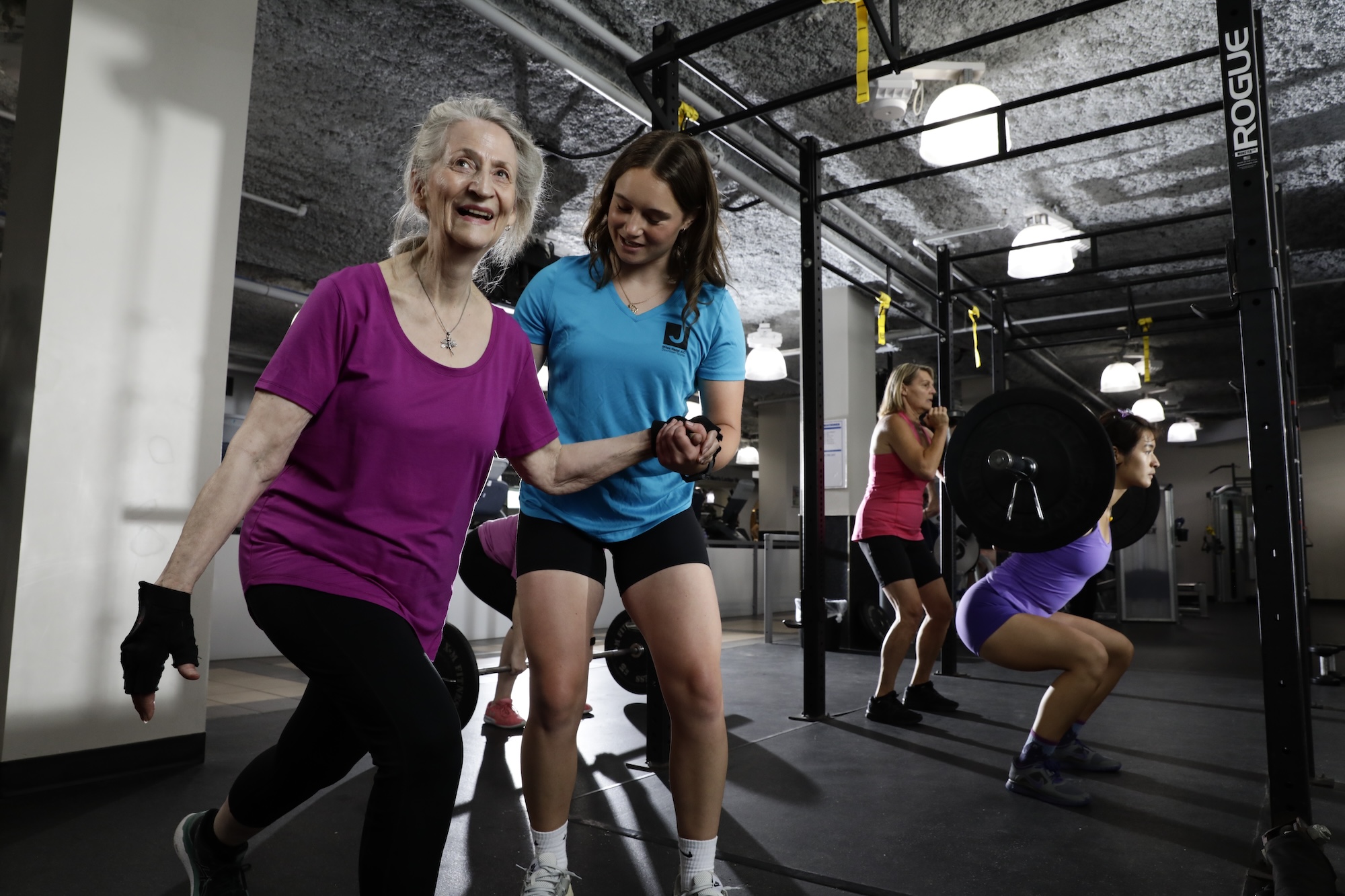 Older woman in purple workout clothes assisted by a trainer in blue during exercise in a gym with others lifting weights in the background.