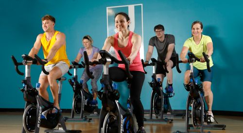 Group of five people smiling and exercising on stationary bikes in a fitness class with a blue wall background.