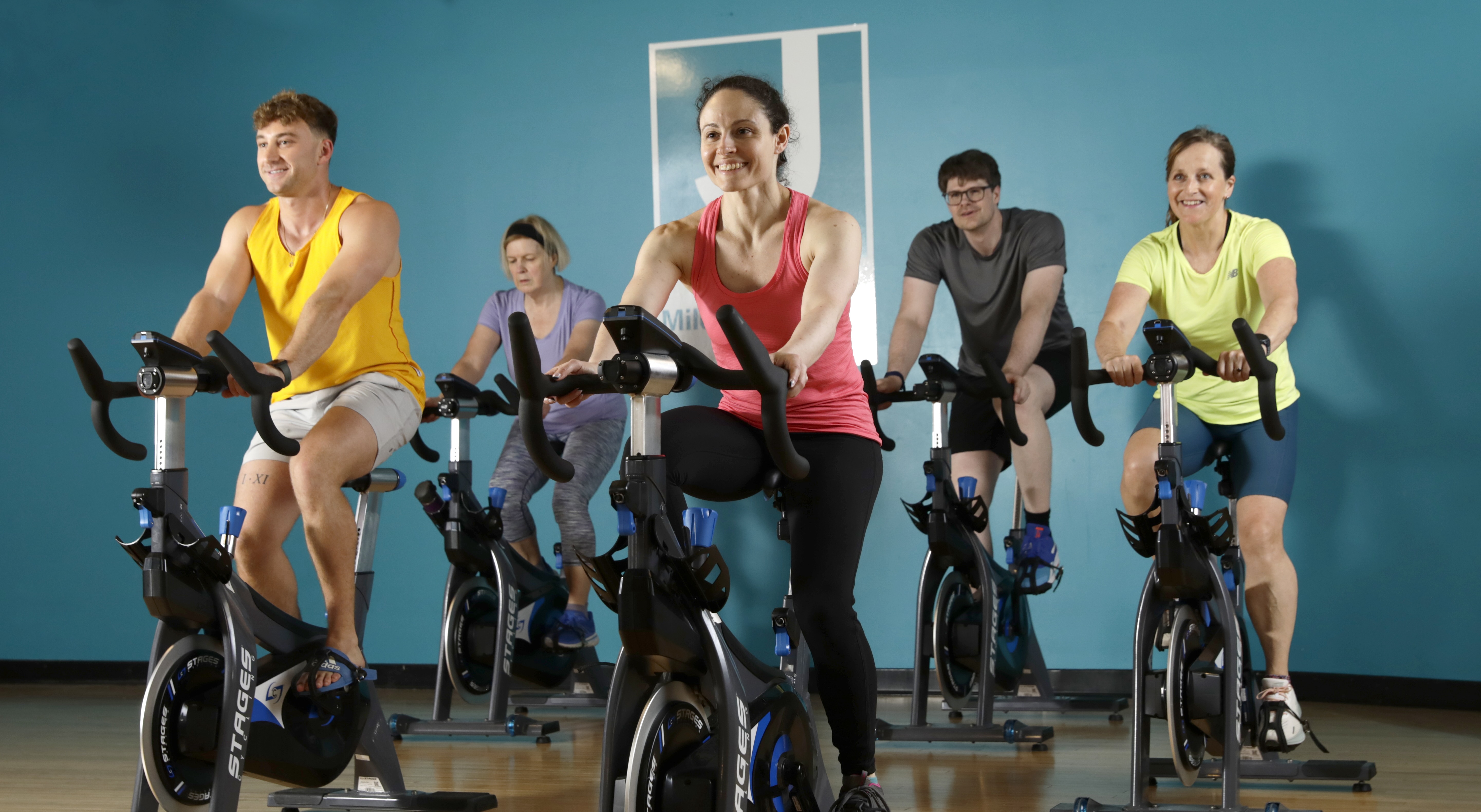 Group of five people smiling and exercising on stationary bikes in a fitness class with a blue wall background.