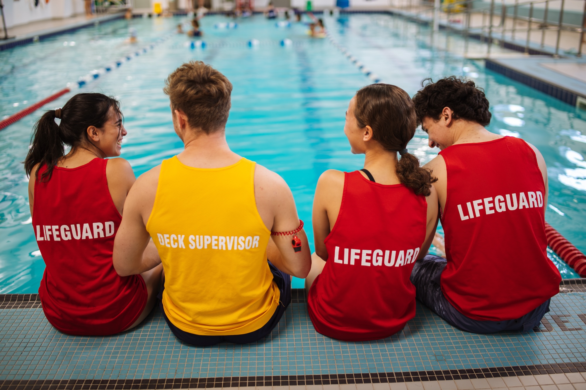Two lifeguards and a deck supervisor sitting by the edge of an indoor swimming pool, facing the water.