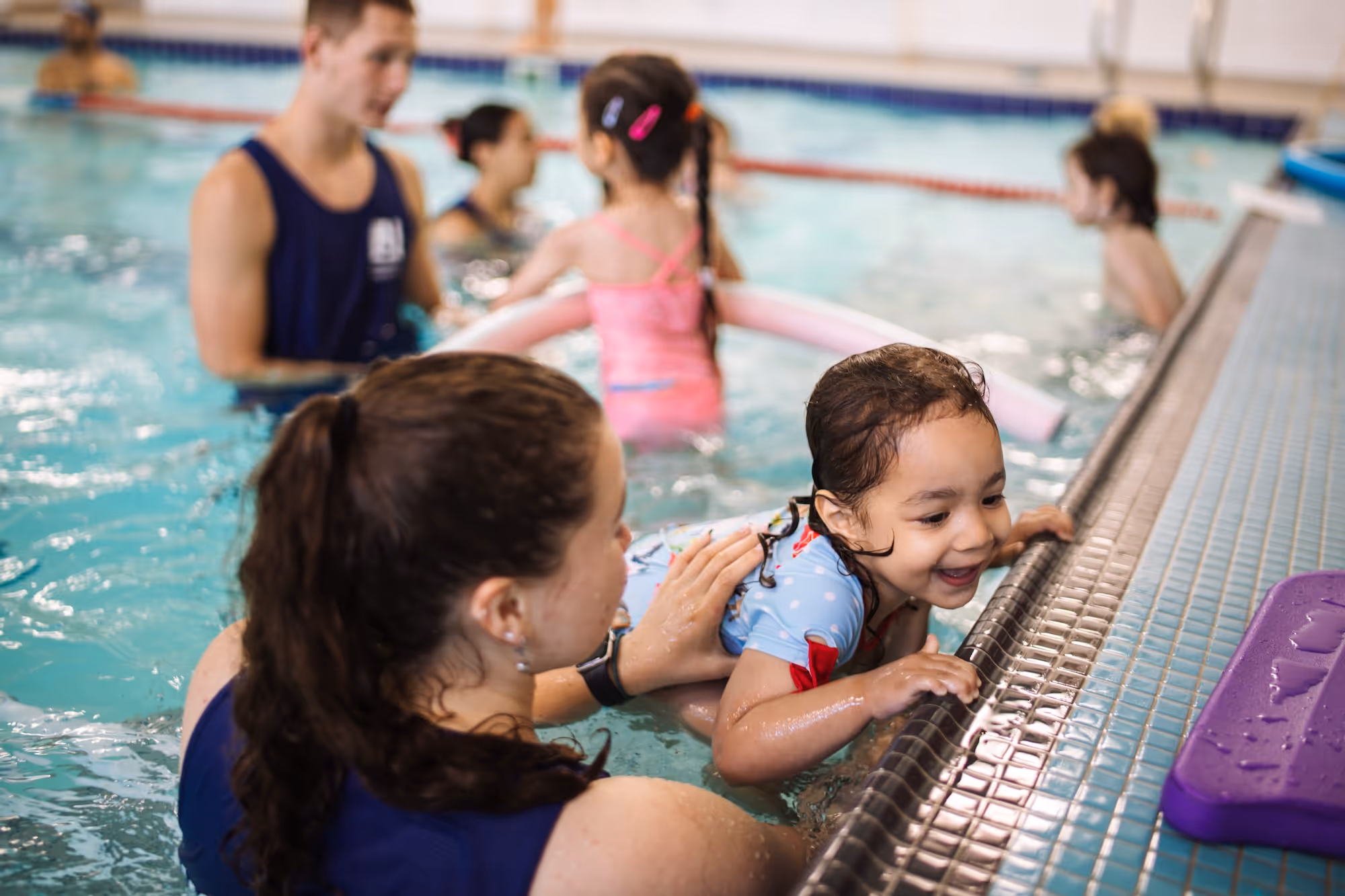 Swimming instructor helps a smiling young girl hold onto the pool edge during a swim lesson with other children and instructors in the background.