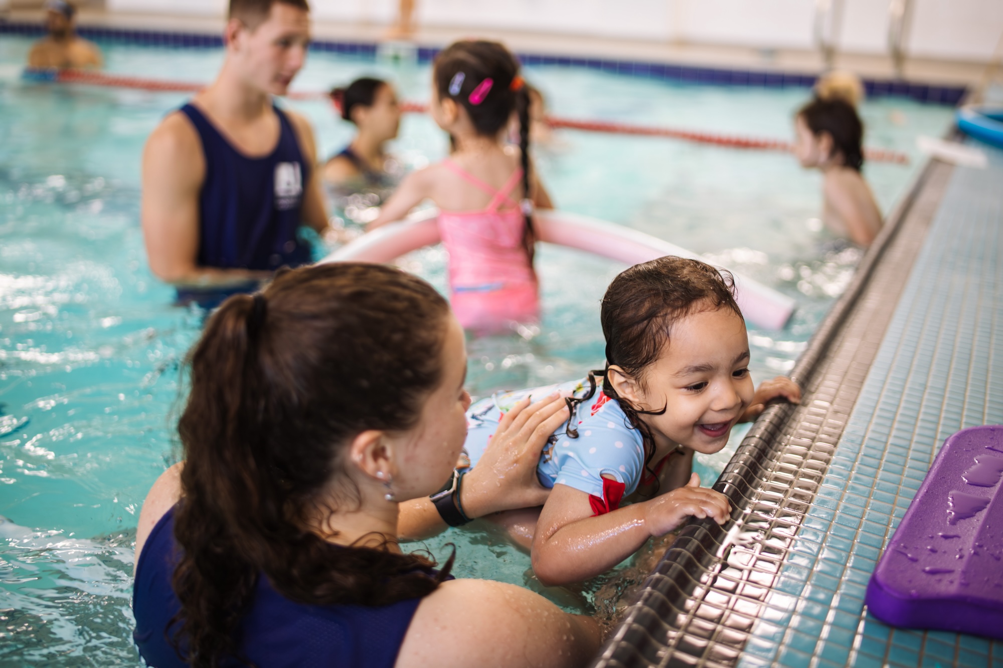 Swimming instructor helps a smiling young girl hold onto the pool edge during a swim lesson with other children and instructors in the background.