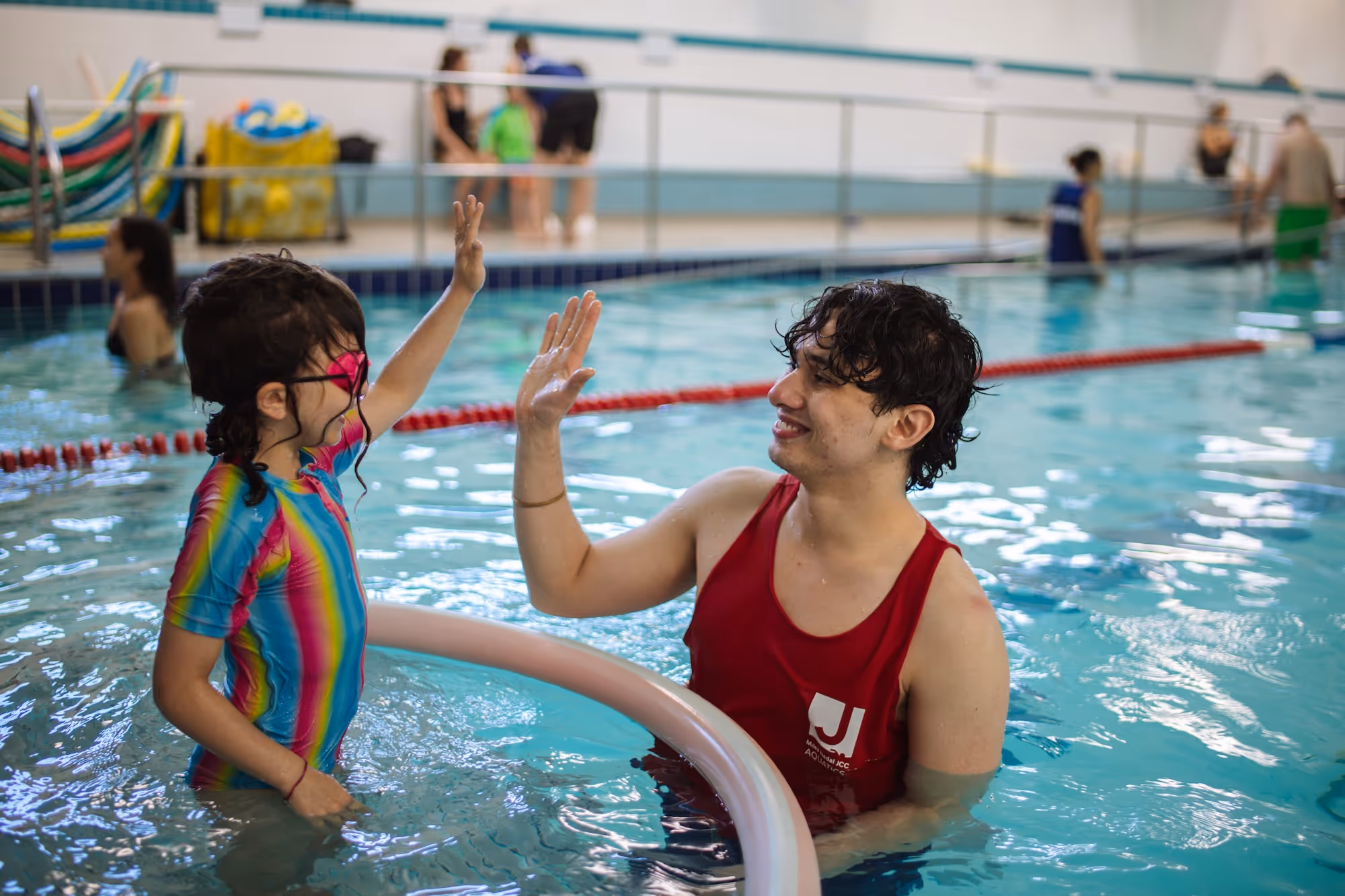 Young girl in colorful swimsuit and pink goggles giving a high-five to a male swim instructor in a red tank top in an indoor pool.