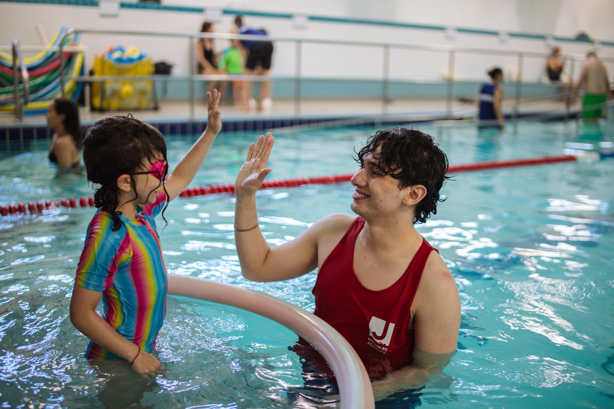 Young girl in colorful swimsuit and pink goggles giving a high-five to a male swim instructor in a red tank top in an indoor pool.