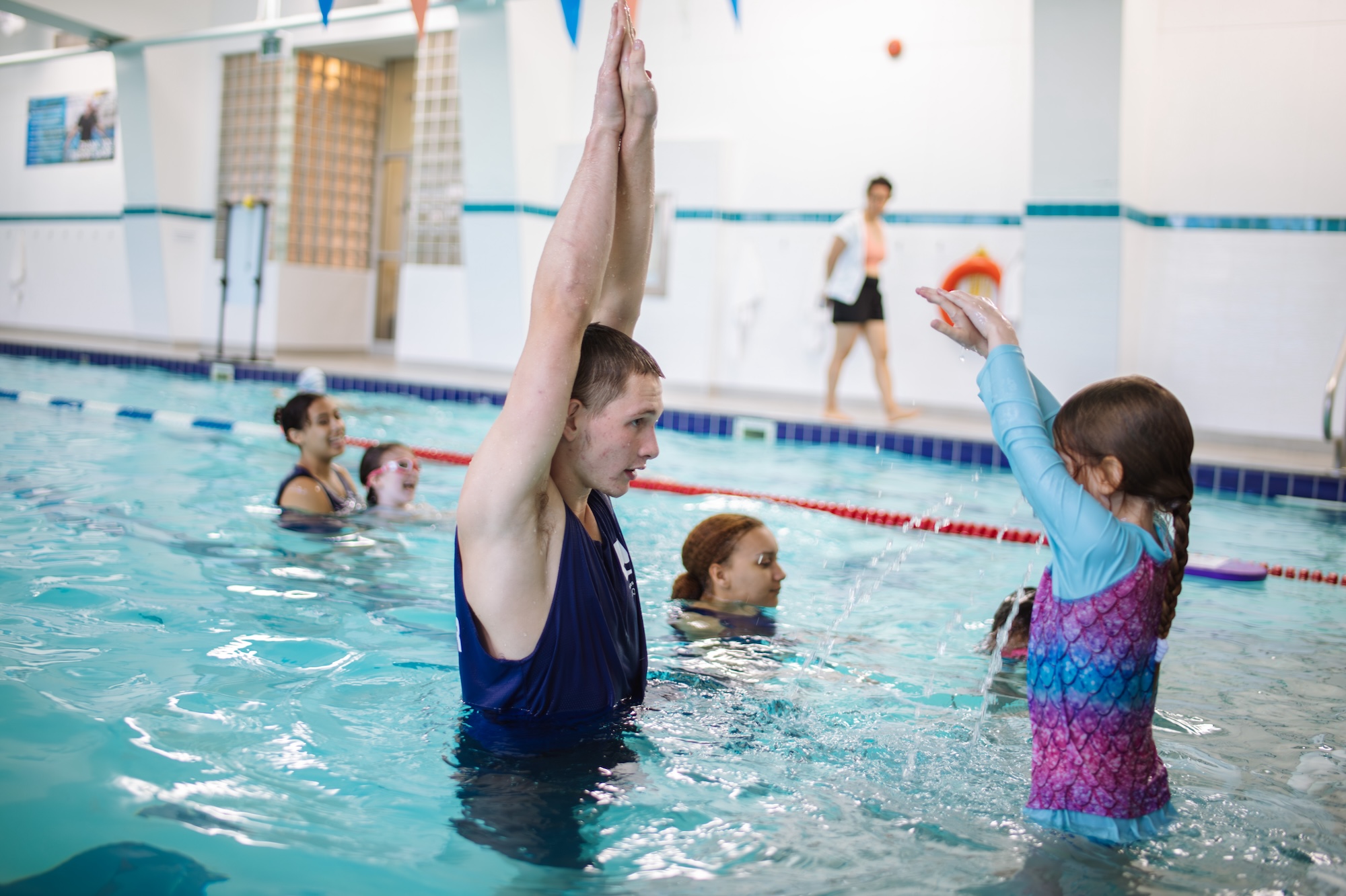 Swimming instructor raising arms in pool teaching young girl in colorful swim shirt, with others in background.