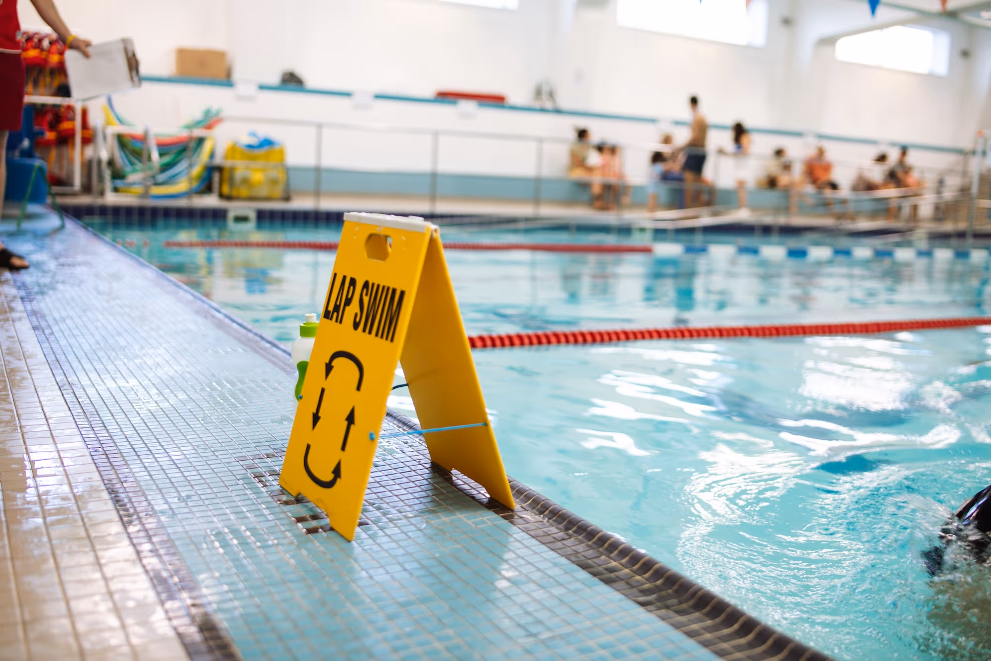 Yellow sign reading 'LAP SWIM' with arrows indicating lap swim direction next to an indoor pool with lane dividers and people in the background.