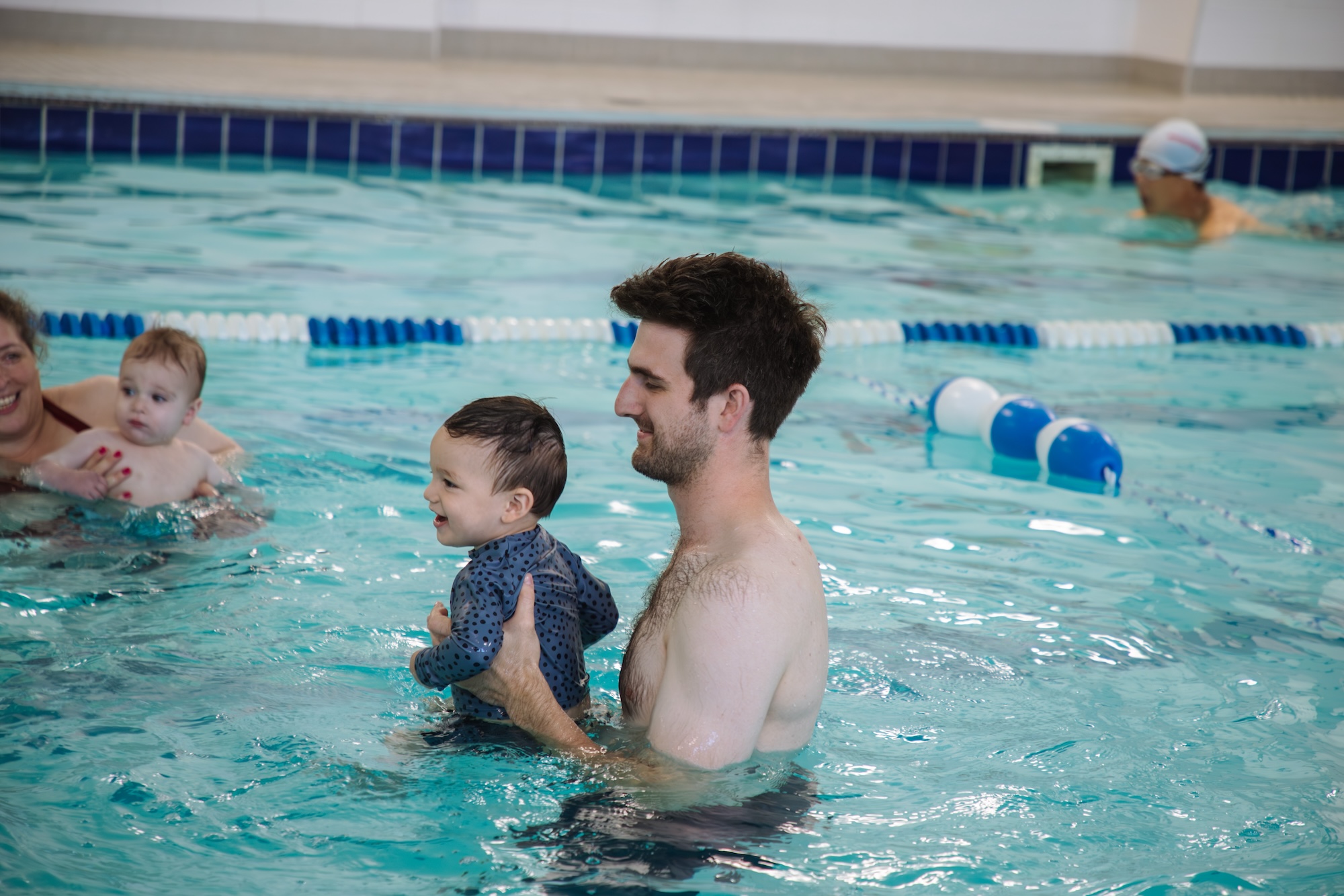 Man holding a smiling toddler in a swimming pool with other adults and children swimming nearby.