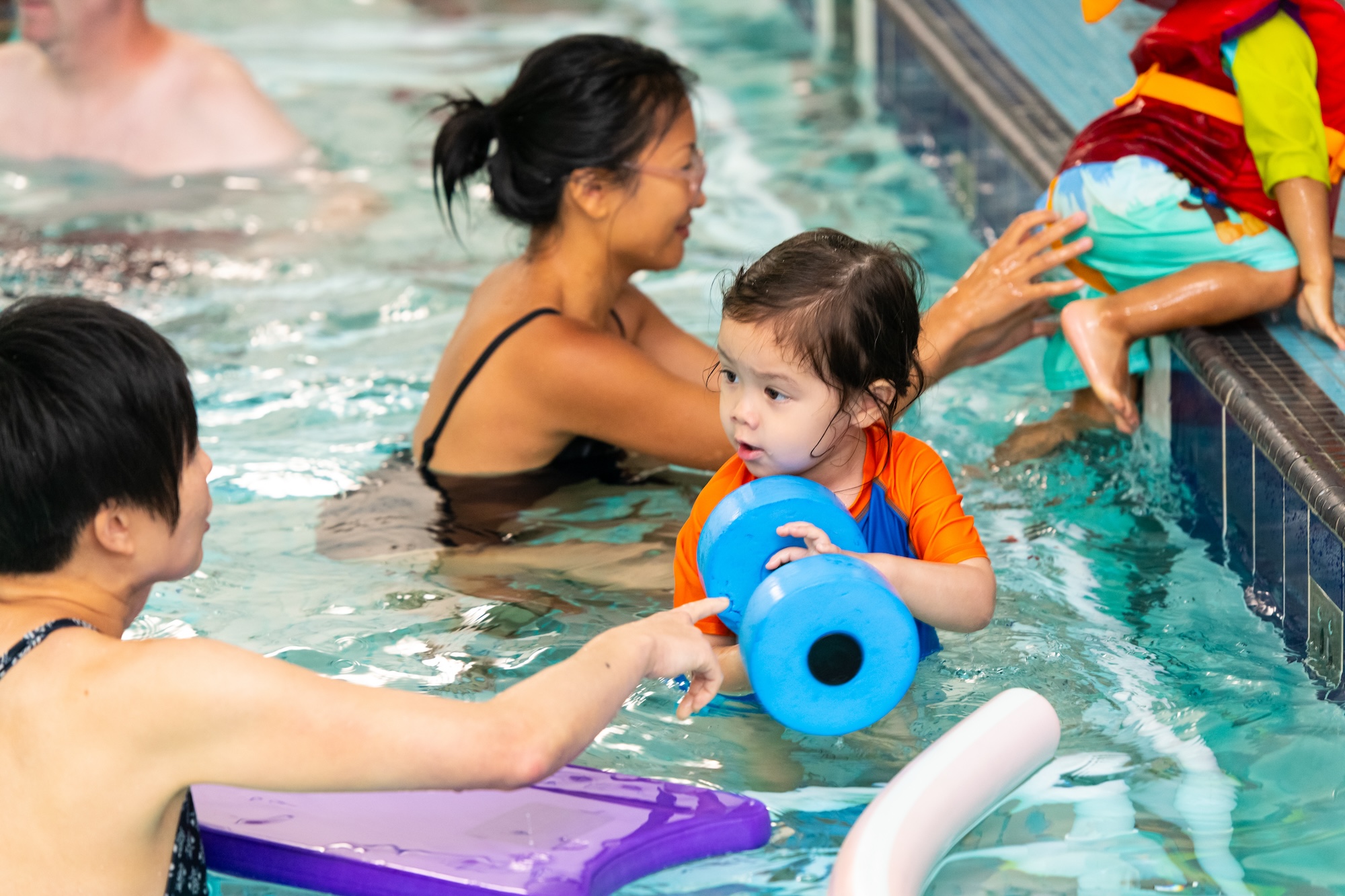 Young child in orange and blue swimwear holding a blue foam dumbbell in a swimming pool during a lesson with an instructor nearby.